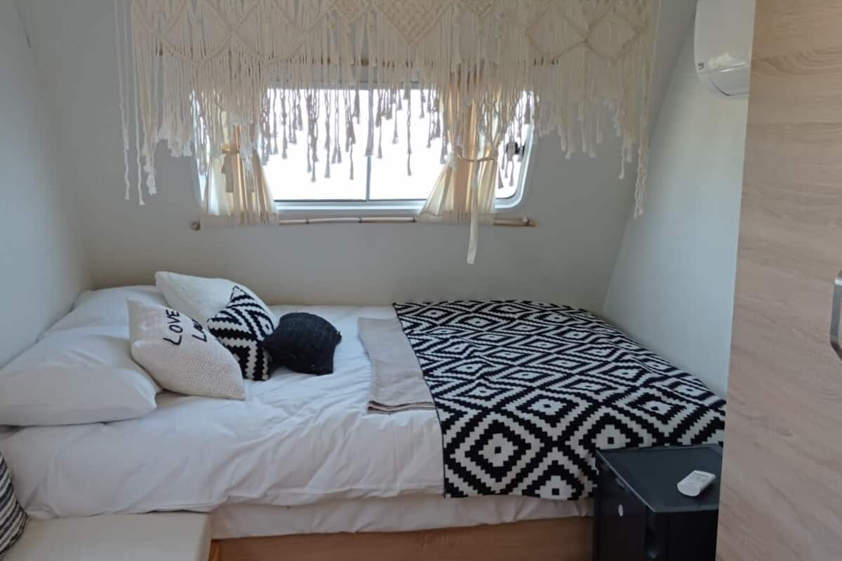 Un dormitorio compacto con cama, una ventana con cortinas de macramé, una mesita de noche pequeña y un armario.