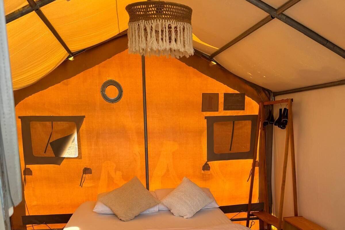 Una habitación tipo tienda de campaña, con cama, lámpara colgante y un pequeño banco de madera.