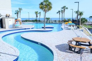A swimming pool with a curved design, surrounded by palm trees and a view of the ocean. There are picnic tables and lounge chairs along the poolside. - 1BR Oceanview at Sand Dunes Resort,Nice High Floor (Myrtle Beach)