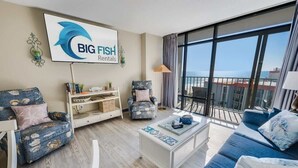 En moderne stue med et stort vindue, en sofa, en lænestol og et sofabord. Et tv viser logoet "BIG FISH Rentals".