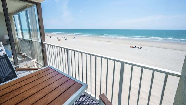 A balcony with a wooden table and chairs overlooking a beach with people and umbrellas. - New Low Floor Direct Ocean Front at Sands Beach Club (Myrtle Beach)