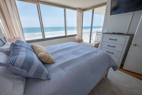 A bedroom with a large bed, a dresser, and a TV. It has a view of the beach and ocean.