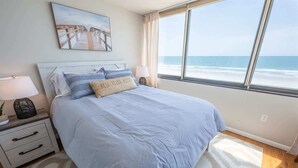 A bedroom with a bed, a nightstand, a lamp, a painting, and a view of the ocean. - New Low Floor Direct Ocean Front at Sands Beach Club (Myrtle Beach)