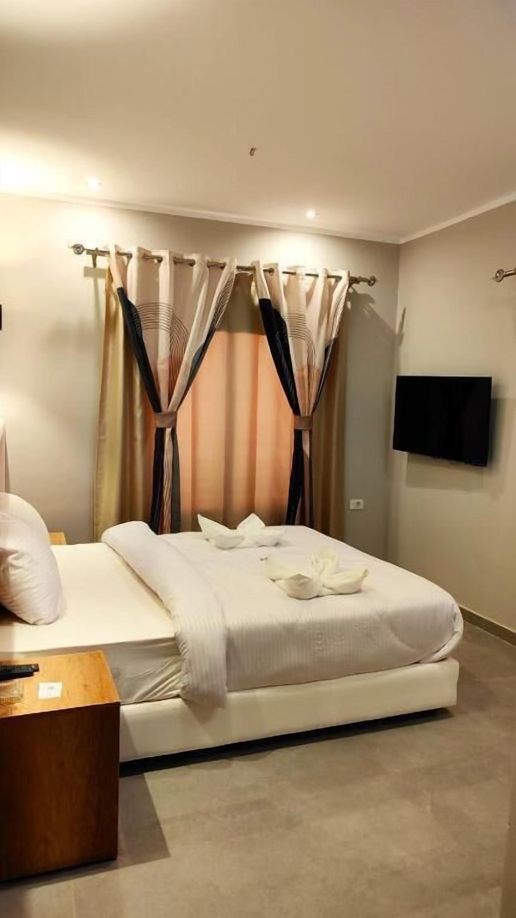 A hotel room with a bed, curtains, a flat-screen TV, and a wooden nightstand.