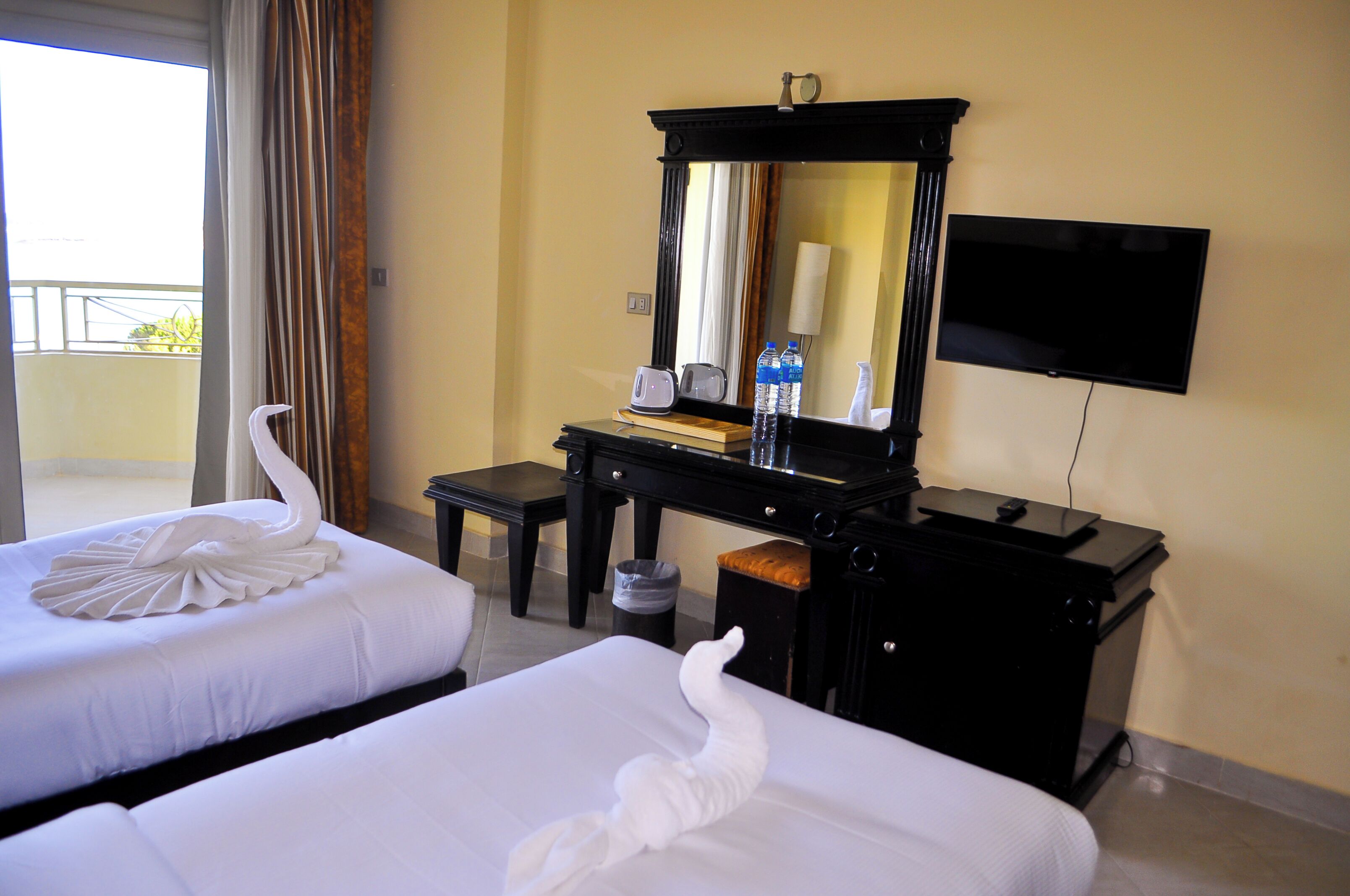 A hotel room with two beds, a desk with a mirror, a television, and a view of the outdoors.