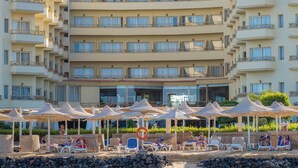 A multi-story hotel with balconies, a pool area with sun loungers and umbrellas, and a view of the surrounding greenery.
