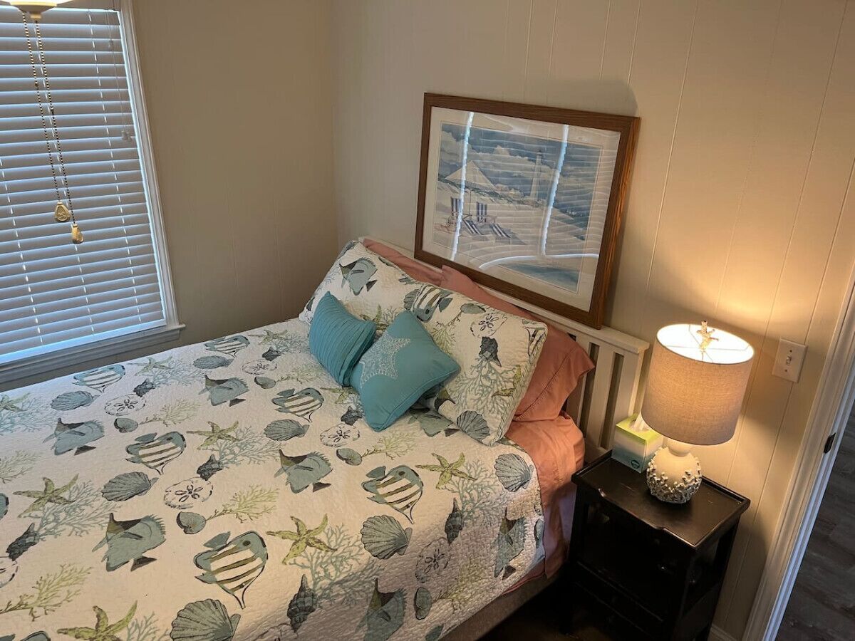 Une chambre à coucher avec un lit, une table de chevet avec une lampe, un tableau encadré au mur et une fenêtre avec des stores.