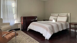 Een slaapkamer met een groot bed, een stoel, een tafeltje en een raam met gordijnen.