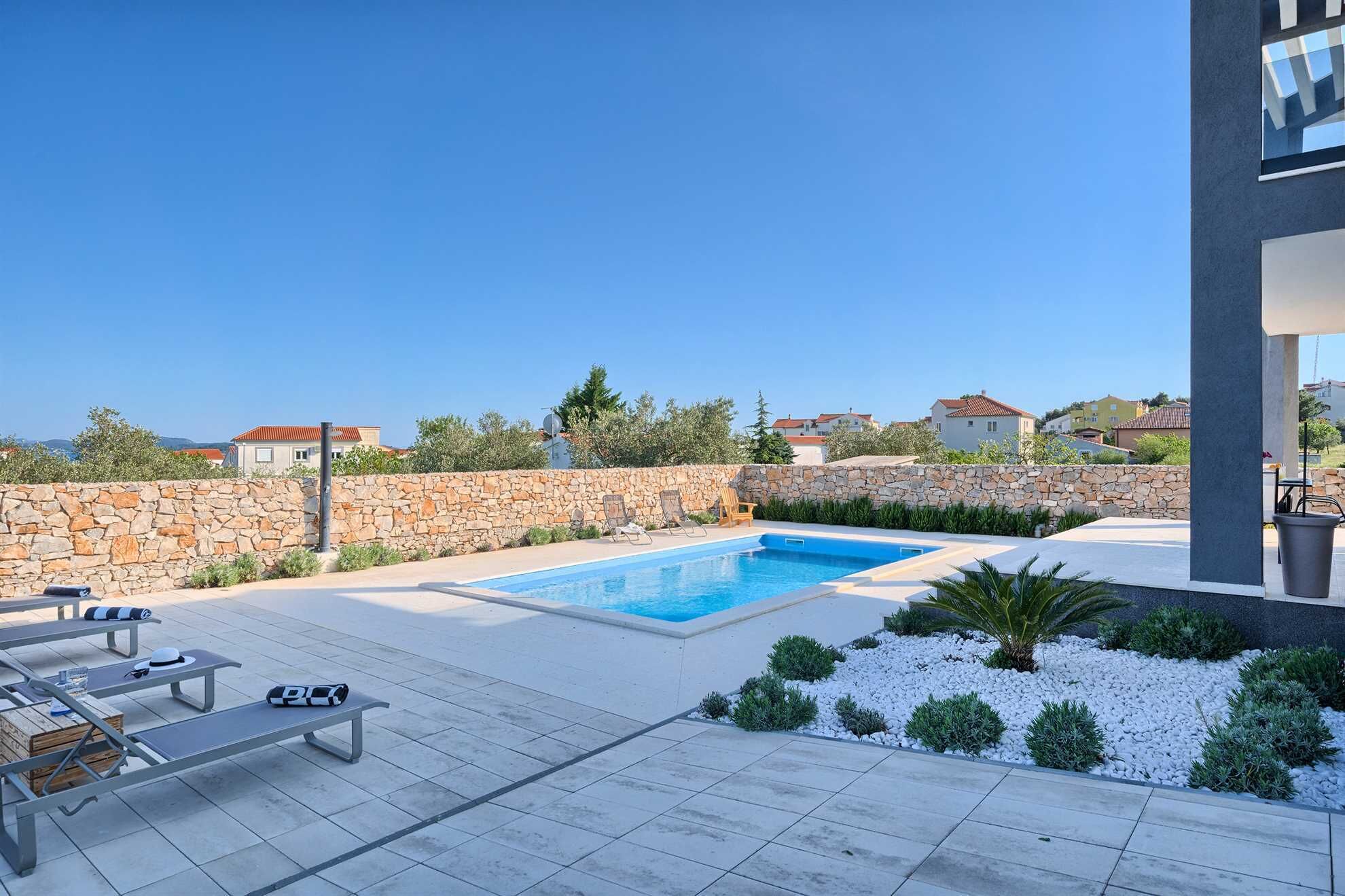 Une piscine entourée de chaises longues et d’un mur de pierre, elle-même bordée d’un jardin aménagé et avec des immeubles d’habitation en arrière-plan.