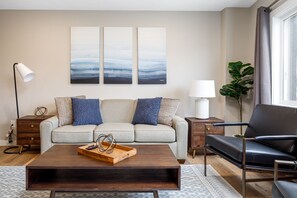 A living room with a sofa, coffee table, and a chair. There are three paintings on the wall, a lamp, and a plant.