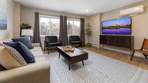 Un salon comprenant un canapé, deux fauteuils, une table basse, un téléviseur à écran plat et une grande fenêtre avec des rideaux.