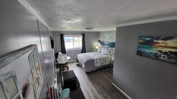 A compact hotel room with a bed, a small desk, a chair, a window, and a painting on the wall.