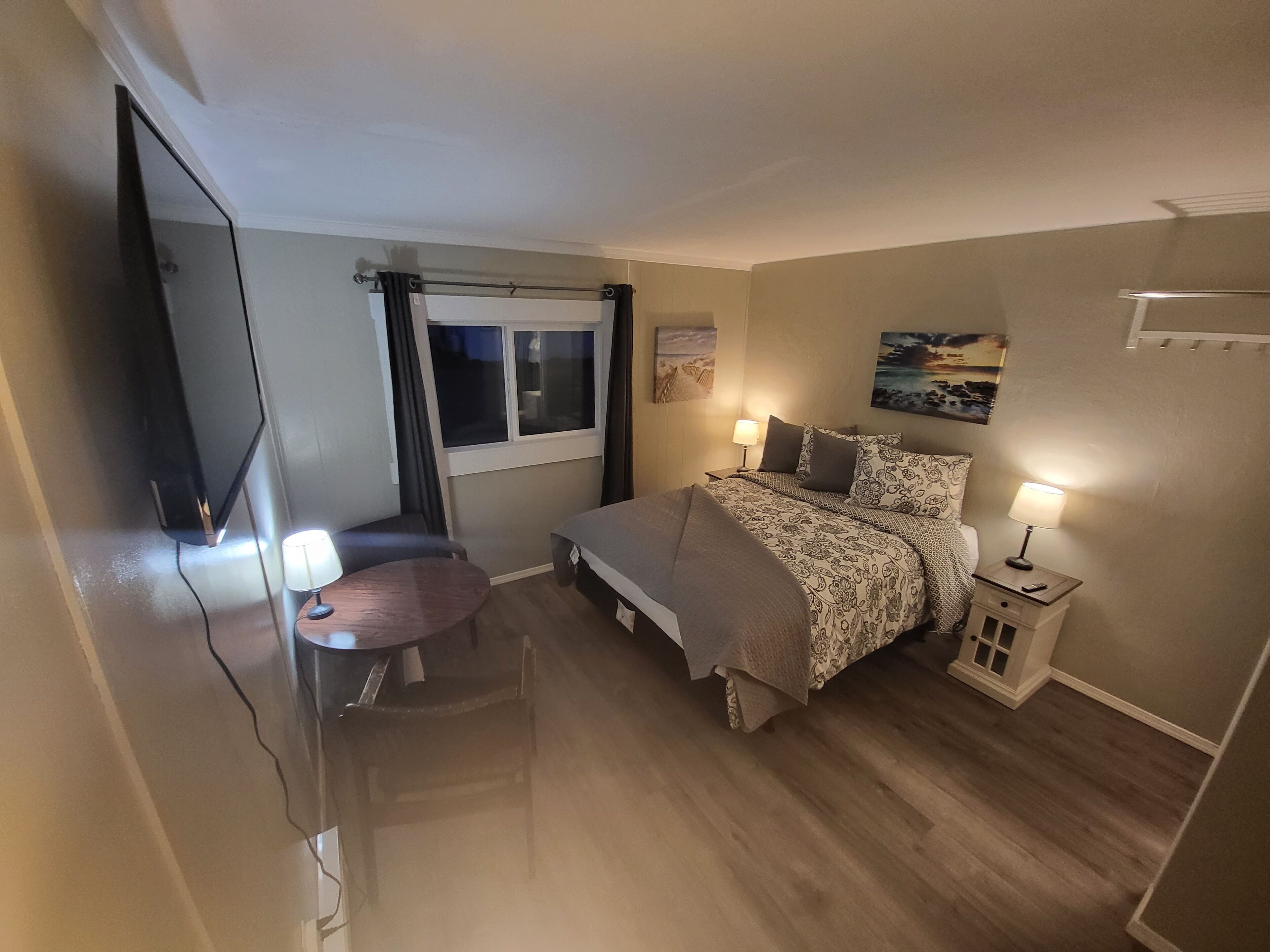 A hotel room with a bed, bedside lamps, a small table, a chair, a TV, and a window with curtains.
