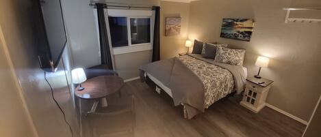A hotel room with a bed, bedside lamps, a small table, a chair, a TV, and a window with curtains.