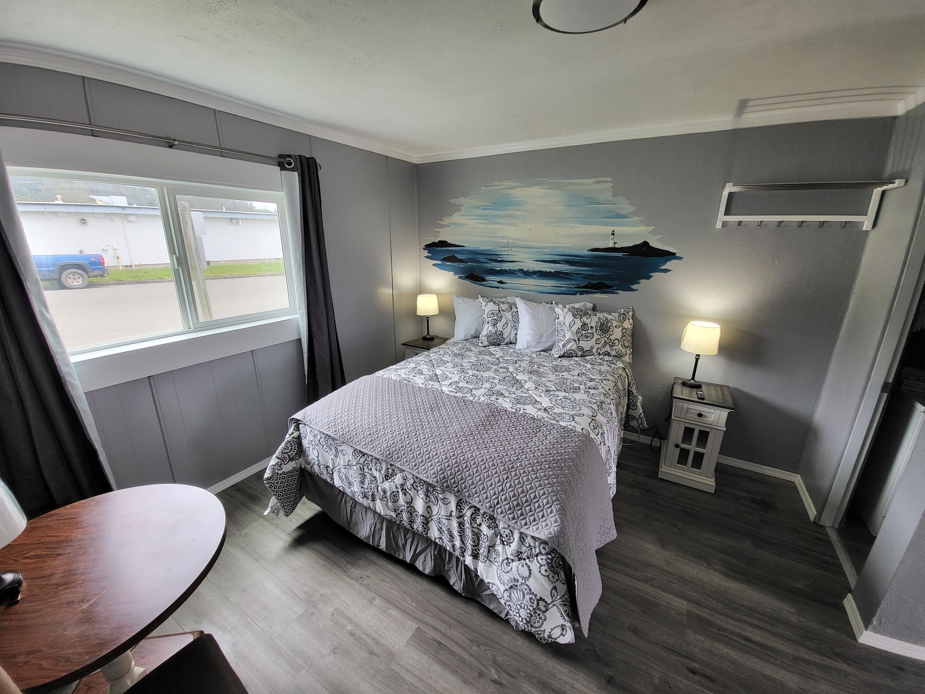 A hotel room with a bed, two bedside lamps, a window with a view of a blue truck, and a wall mural.