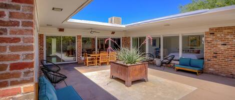 A modern patio with a brick wall, a wooden bench, a potted plant, and a dining area with a table and chairs.