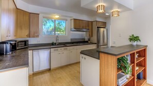 A modern kitchen with wooden cabinets, a built-in oven, a refrigerator, and a microwave. There is a window providing natural light and a potted plant on the counter.