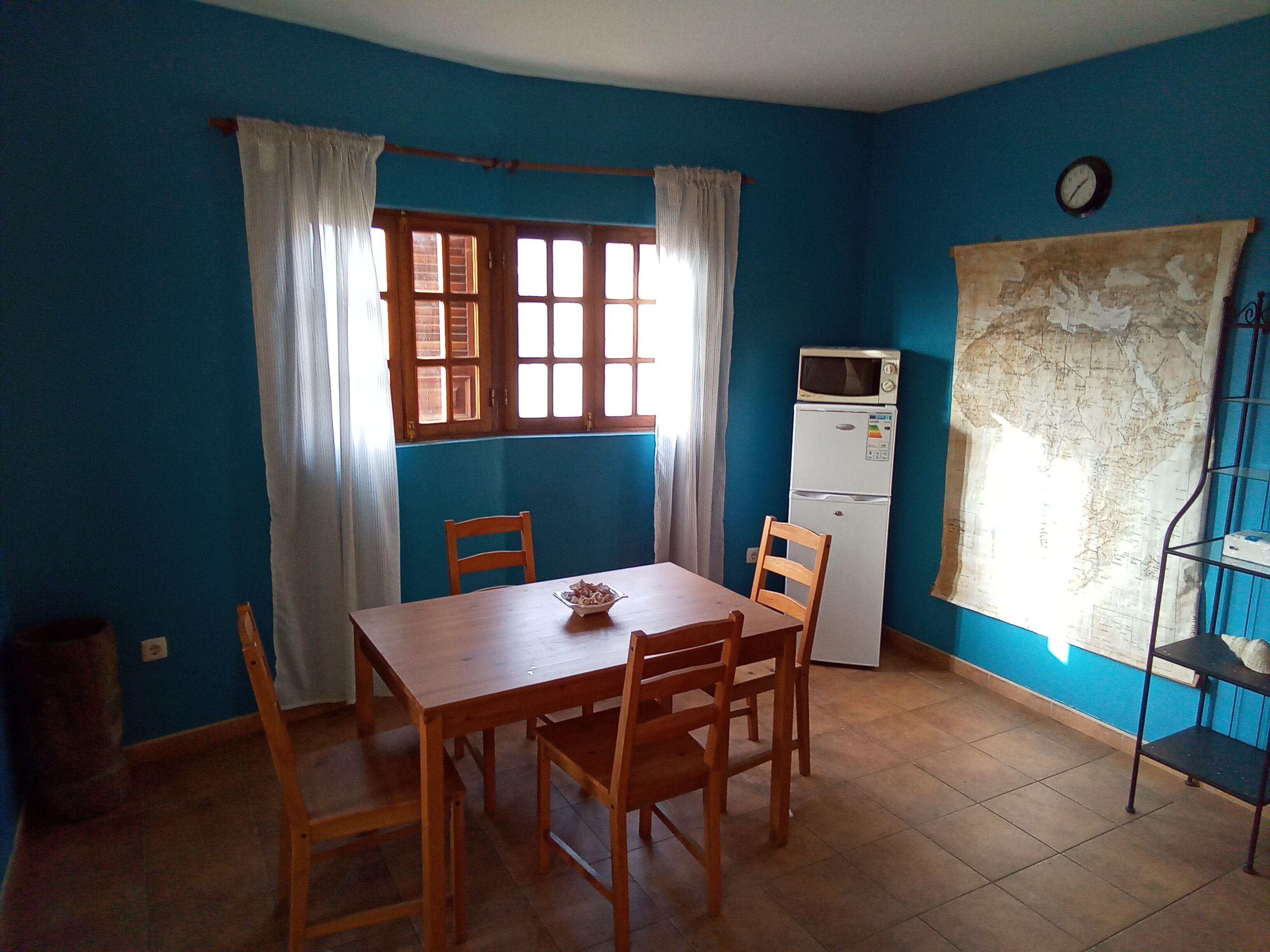 A dining area with a wooden table and chairs, a small refrigerator, a window with curtains, and a wall clock.
