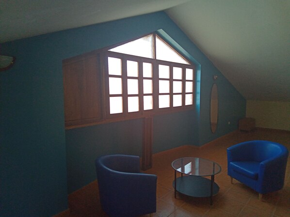 A room with a sloped ceiling, a large window with multiple panes, two blue armchairs, and a small round table. - GREEN SEA APARTMENTS (Tarrafal)