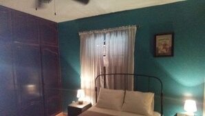 A bedroom with teal walls, a bed with white linens, two bedside tables with lamps, a ceiling fan, and a window with curtains.
