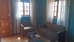 A room with blue walls, a wooden door, two wicker sofas, a glass coffee table, and two windows with white curtains.