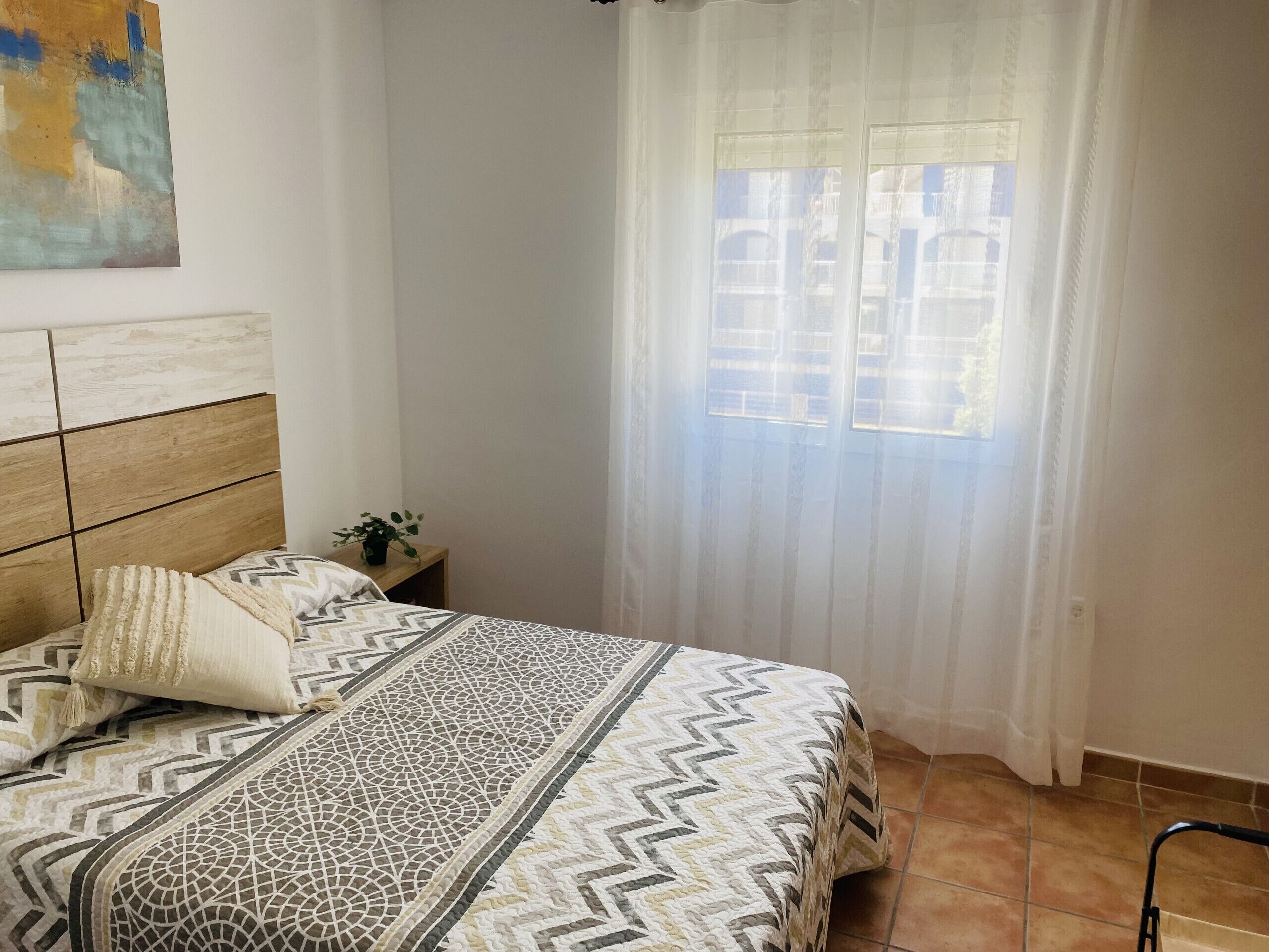 A bedroom with a bed, a headboard, a nightstand, a window with curtains, and a view of buildings outside.