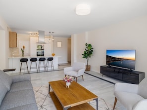 Una sala de estar moderna con un sofá, una mesa de centro y un televisor. Cuenta con una zona de cocina con barra y taburetes.