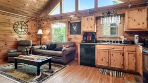 A cozy cabin interior with a wooden ceiling, a dining area with a table and chairs, a kitchen with wooden cabinets, and a large window providing natural light. - Couples Cabin - Private Hot Tub - 1 Mile to Parkway (Pigeon Forge)