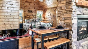 A cozy cabin interior with a wooden dining table, bench seating, a fireplace, and a bed with a window view. - Couples Cabin - Private Hot Tub - 1 Mile to Parkway (Pigeon Forge)