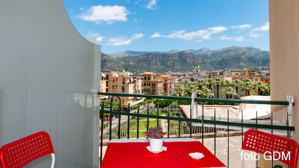 A balcony with a red table and chairs, a potted plant, and a view of a cityscape with mountains in the background. - Casa Christal (Sant´Agnello)