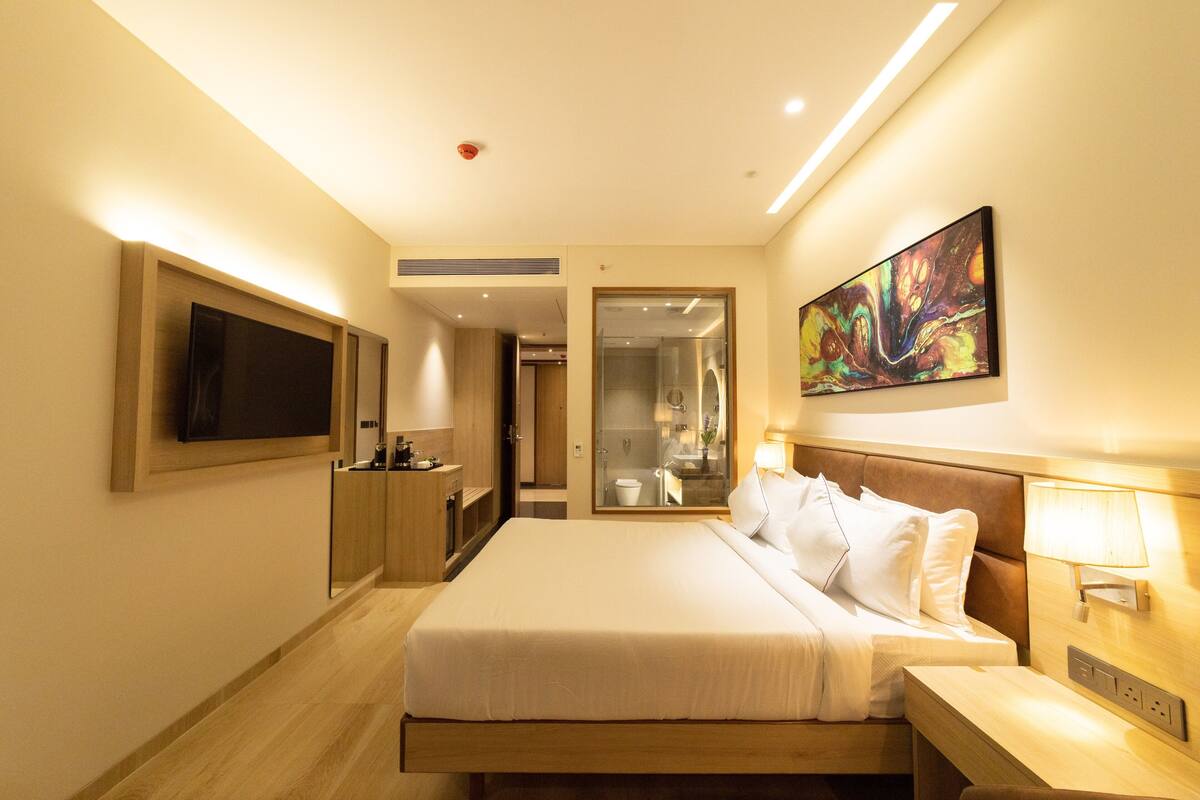 Habitació d'hotel moderna amb un llit gran, un televisor de pantalla plana fixat a la paret, una taula de nit amb una làmpada i un bany visible a través d'una porta oberta.