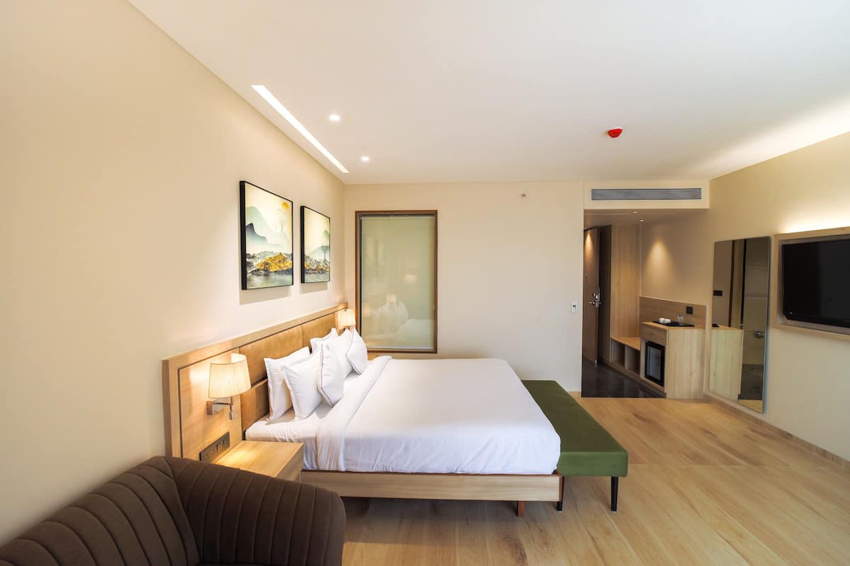 Habitació d'hotel moderna amb un llit gran, una taula de nit amb una làmpada, una banca verda, un televisor i una cuineta.