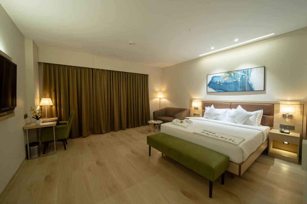 Habitació d'hotel amb llit, escriptori, cadira, banc i una finestra gran amb cortines.