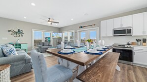 A modern kitchen with white cabinets, a wooden dining table set for four, and a large window with blinds. - Private beach/Luxury home/Private pool (Gulf Shores)