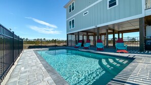 A swimming pool with a stone deck, a covered seating area with Adirondack chairs, and a two-story building with a flat roof. - Private beach/Luxury home/Private pool (Gulf Shores)