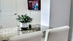 A dining area with a granite table, white chairs, a potted plant, and a wall-mounted TV. - Amazing Melly's (Tampa)