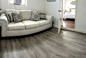 A living room with a beige sofa, patterned cushions, and a wooden floor. A door leads to a bedroom with a bed and a nightstand. - Amazing Melly's (Tampa)