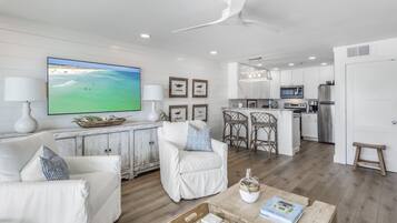 A modern living room with a large TV, white furniture, and a kitchen in the background.