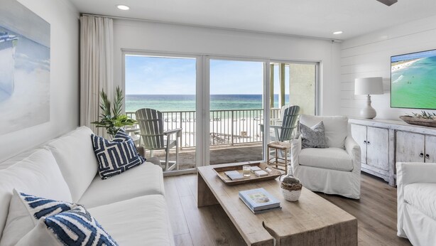 Een ruime woonkamer met een witte bank, fauteuils en een salontafel. Een groot raam biedt uitzicht op het strand en de zee.