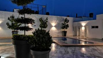 Zona de piscina al aire libre con plantas en macetas y una pared blanca con un accesorio de iluminación.