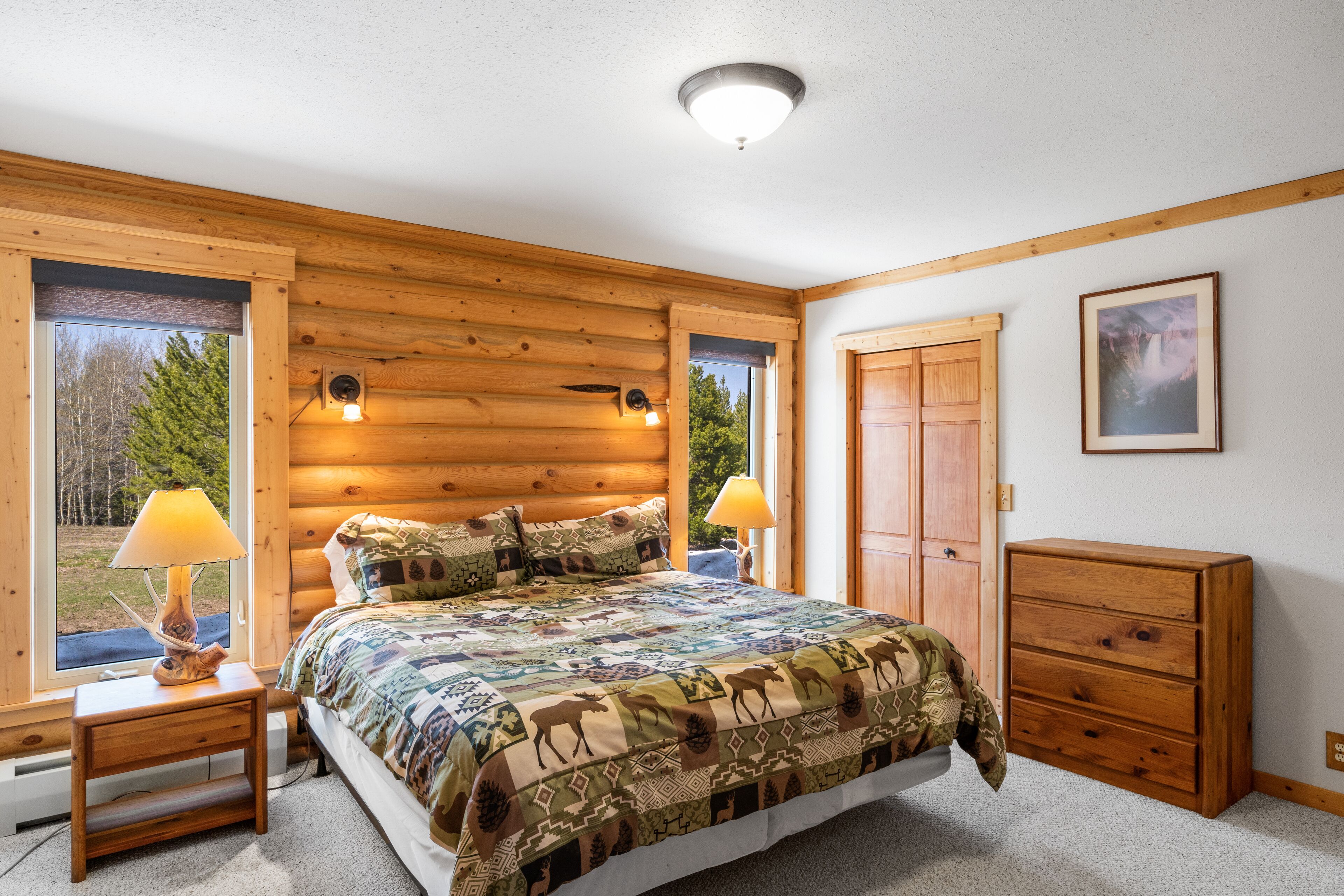 Ein Schlafzimmer in einer Blockhütte mit einem Bett, zwei Nachttischen mit Lampen, einer Kommode und einem gerahmten Bild an der Wand.