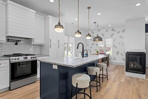 A modern kitchen with white cabinetry, a central island with stools, pendant lighting, and a fireplace. - Mountain Home near Lakes w/Hot Tub & Putting Green (Heber City)