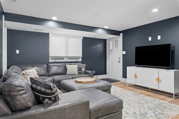 A modern living room with a grey sectional sofa, a flat-screen TV on a white cabinet, and a large window with blinds. - Mountain Home near Lakes w/Hot Tub & Putting Green (Heber City)