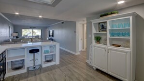 A modern kitchen with a central island, bar stools, and a built-in cabinet with decorative items. - El Matador 528 - FREE TICKETS!! Your stay includes free tickets to many area attractions! (Fort Walton Beach)