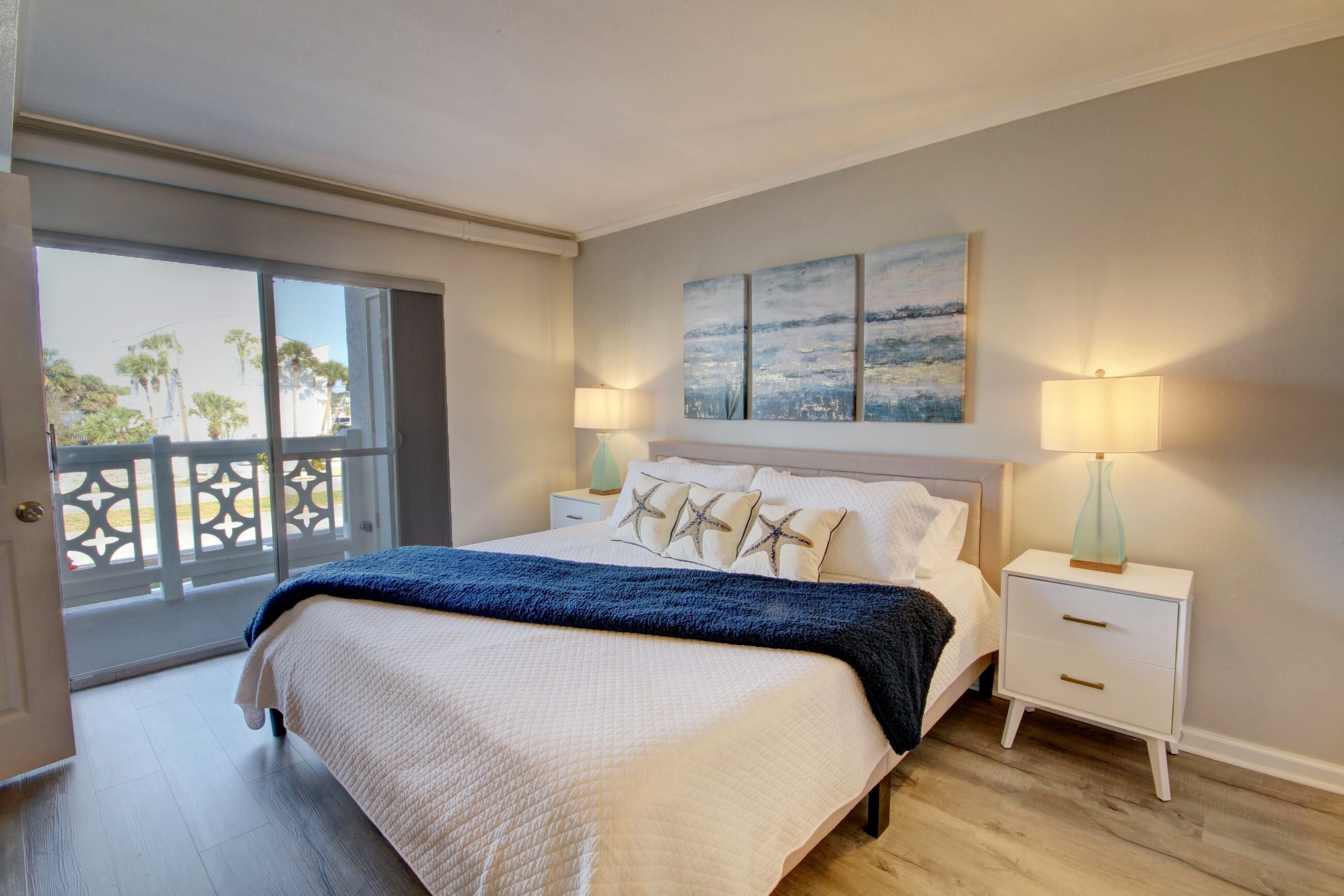 Un dormitorio con una cama grande, dos mesitas de noche con lámparas, vista a través de una puerta corredera y un cuadro en la pared.