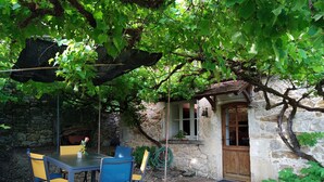 A rustic stone building with a wooden door, a small window, and a table with blue and yellow chairs. - Quercy house at the foot of the cliffs (Vayrac)