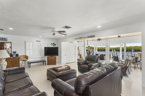 A spacious living area with a television, leather sofas, a dining area with outdoor seating, and a view of a body of water.