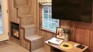 Een gezellige kamer met houten inrichting, een open haard, een aan de muur bevestigde televisie en een klein tafeltje met een fotolijst en een vaas.
