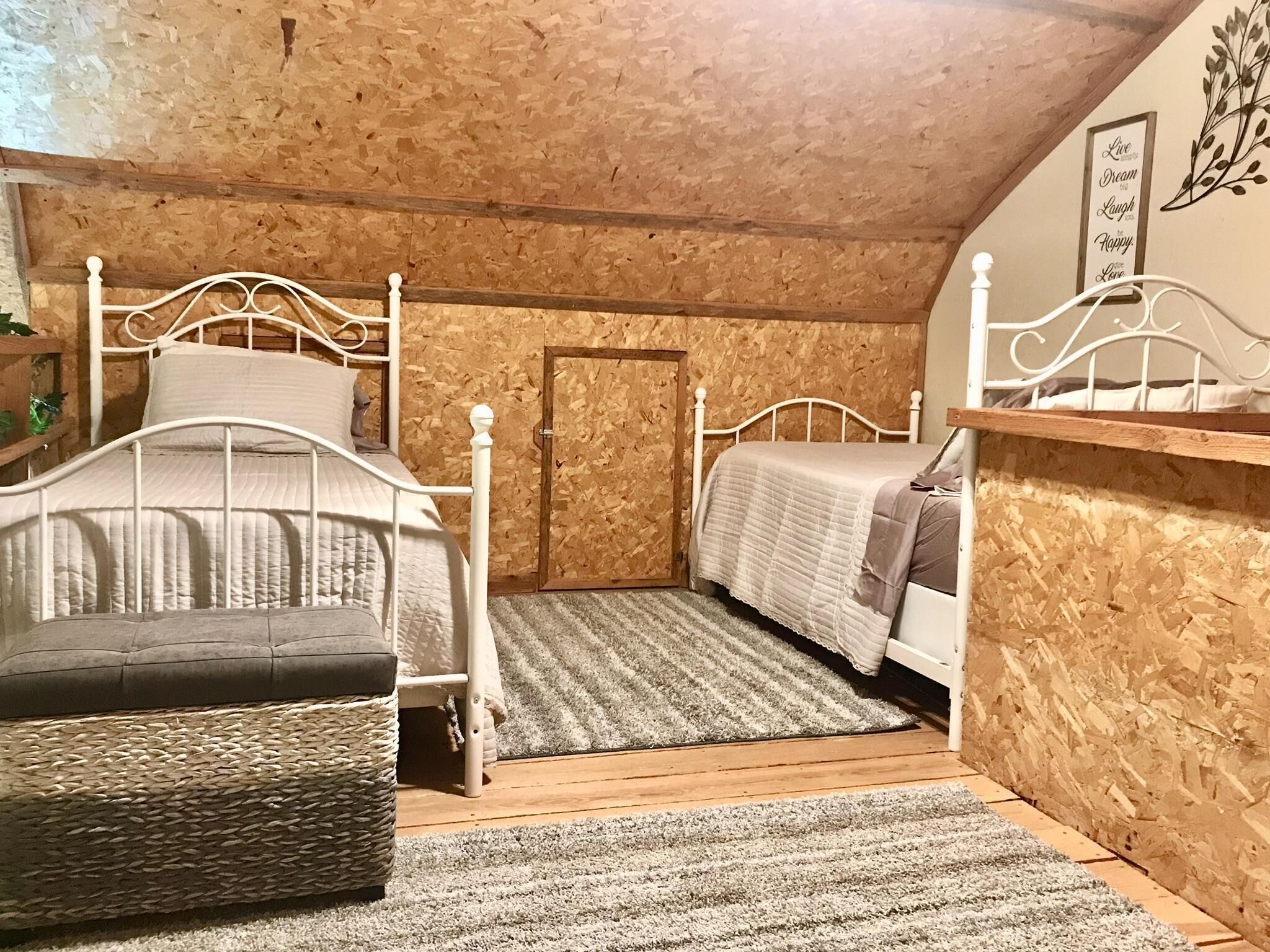 Quarto com duas camas de metal branco, um banco de fibra natural, piso de madeira e parede com uma placa decorativa.