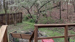 Une terrasse en bois avec un banc et un tabouret rouge, entourée d’une clôture en bois et d’arbres aux fleurs blanches.